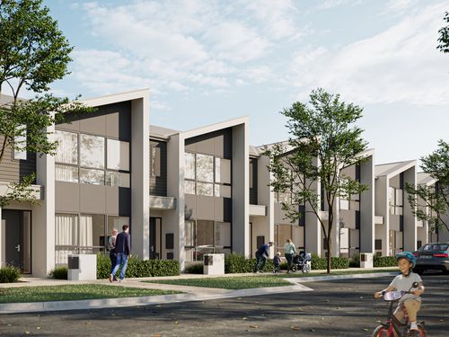 A row of modern townhomes at Riverlea along a quiet street on a bright, clear day. Pedestrians are walking along the footpath while a child is riding a bicycle along the edge of road.