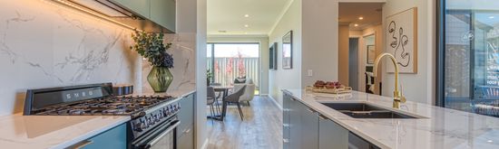 The modern kitchen and adjoining living space of a home at the Riverlea Display World, with marble benchtops and modern, minimalistic fittings and abstract artwork adorning the walls of the living area and nearby hallway.