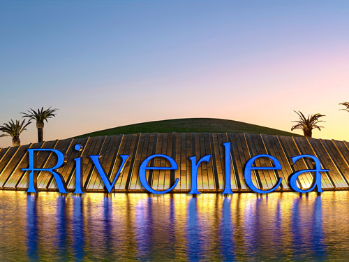 The entry at Riverlea, featuring a large lake, palm trees in the background, and a large sign displaying the Riverlea Logo