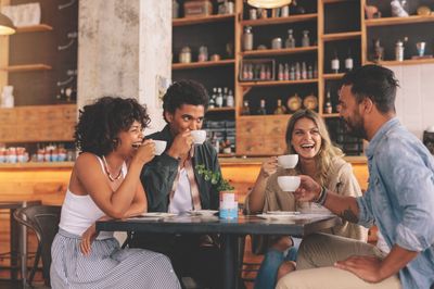 A group of young people laughing and drinking coffee while sitting around a table in a modern coffee shop.