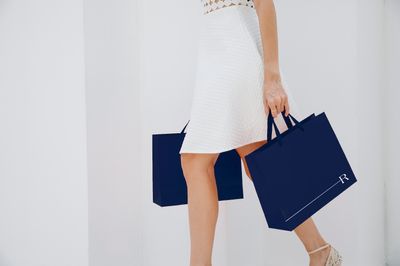 A person wearing a bright white dress carrying two navy coloured paper shopping bags with the Riverlea logo on them