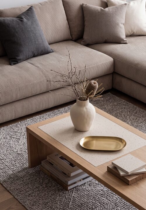 A ceramic vase with dried native branches and a brass tray sit on a timber-framed coffee table beside a corduroy sectional sofa with charcoal and neutral cushions, on a textured wool rug.