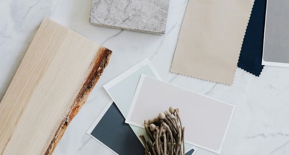 A flat lay of interior finishes on a marble benchtop, including a timber veneer sample, a stone slab, fabric swatches and paint colour cards.