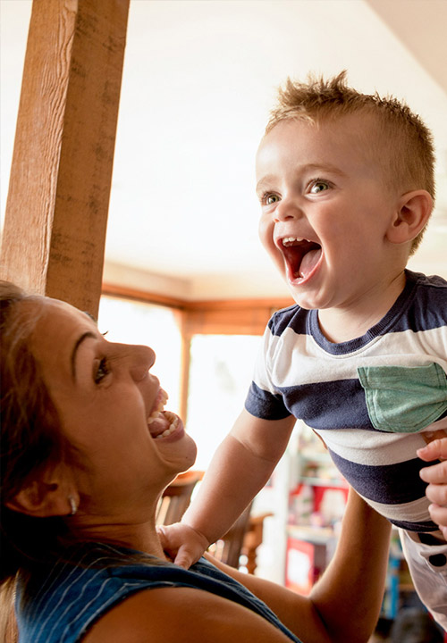 A woman smiles as she lifts a laughing baby in a striped shirt inside a sunlit room.