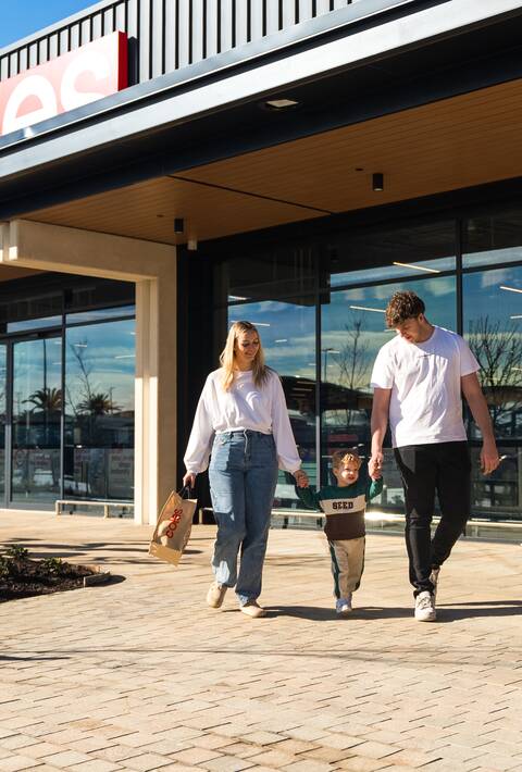 A smiling couple walking while holding their young child’s hands leaving a new Coles supermarket.