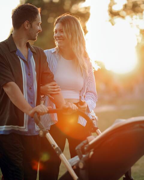A couple pushing a pram alongside a calm lake as the sun begins to set in the distance.