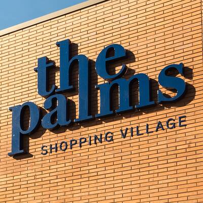 A sign for The Palms Shopping Village in deep blue on a tall brown brick wall.