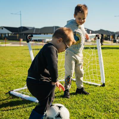 Two young children laughing while playing with a soccer ball. One child is defending a small goal while the other kicks the ball towards it.
