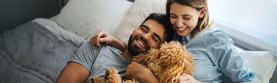 A smiling couple lounging on a bed while hugging a small, furry dog.