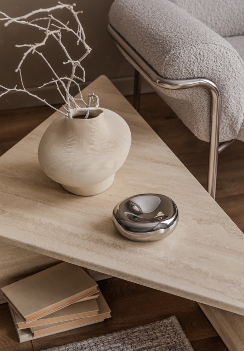 A textured vase with bare branches and a reflective metallic décor piece sit on a stone coffee table beside a cosy upholstered chair.